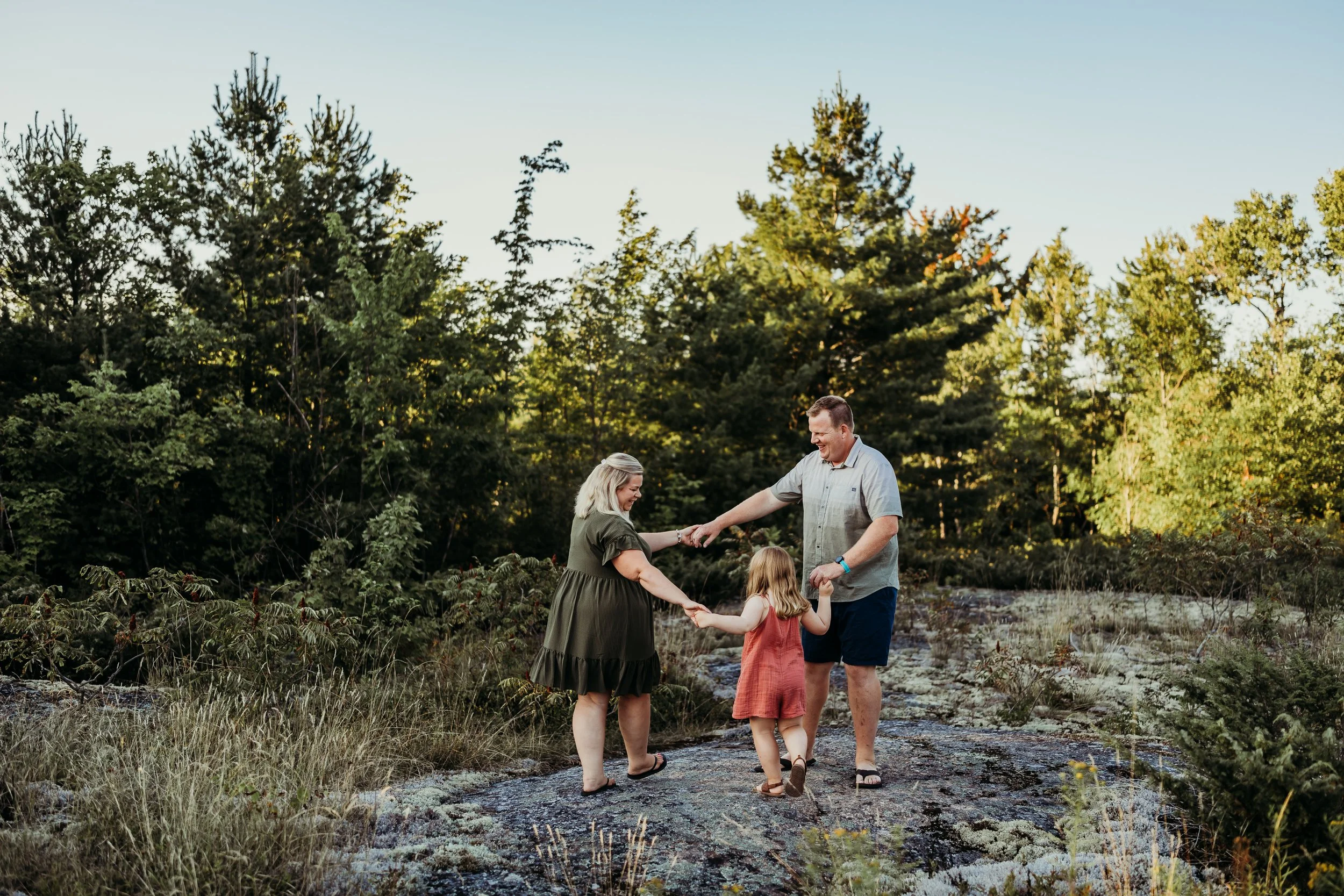 A family of three holding hands and dancing outdoors on a rocky area surrounded by trees, with a clear sky.