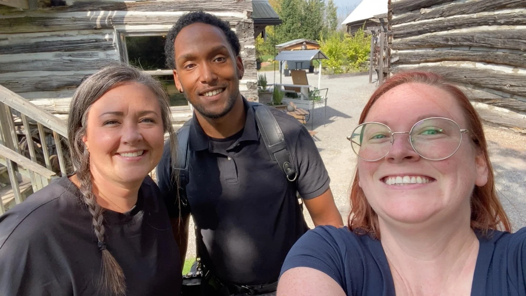 Three friends smiling and taking a selfie outdoors in front of rustic wooden buildings, with trees and a clear sky in the background.