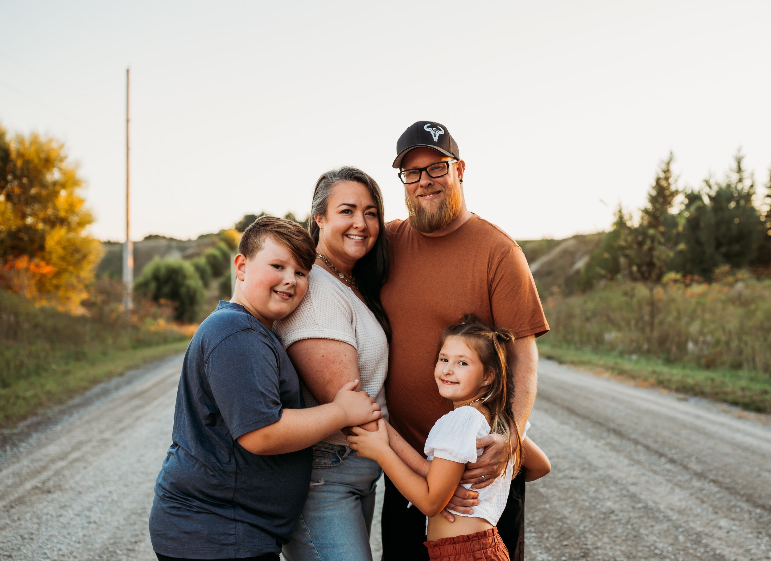 Family of five standing on a dirt road outdoors, smiling, with trees and a sunset background.