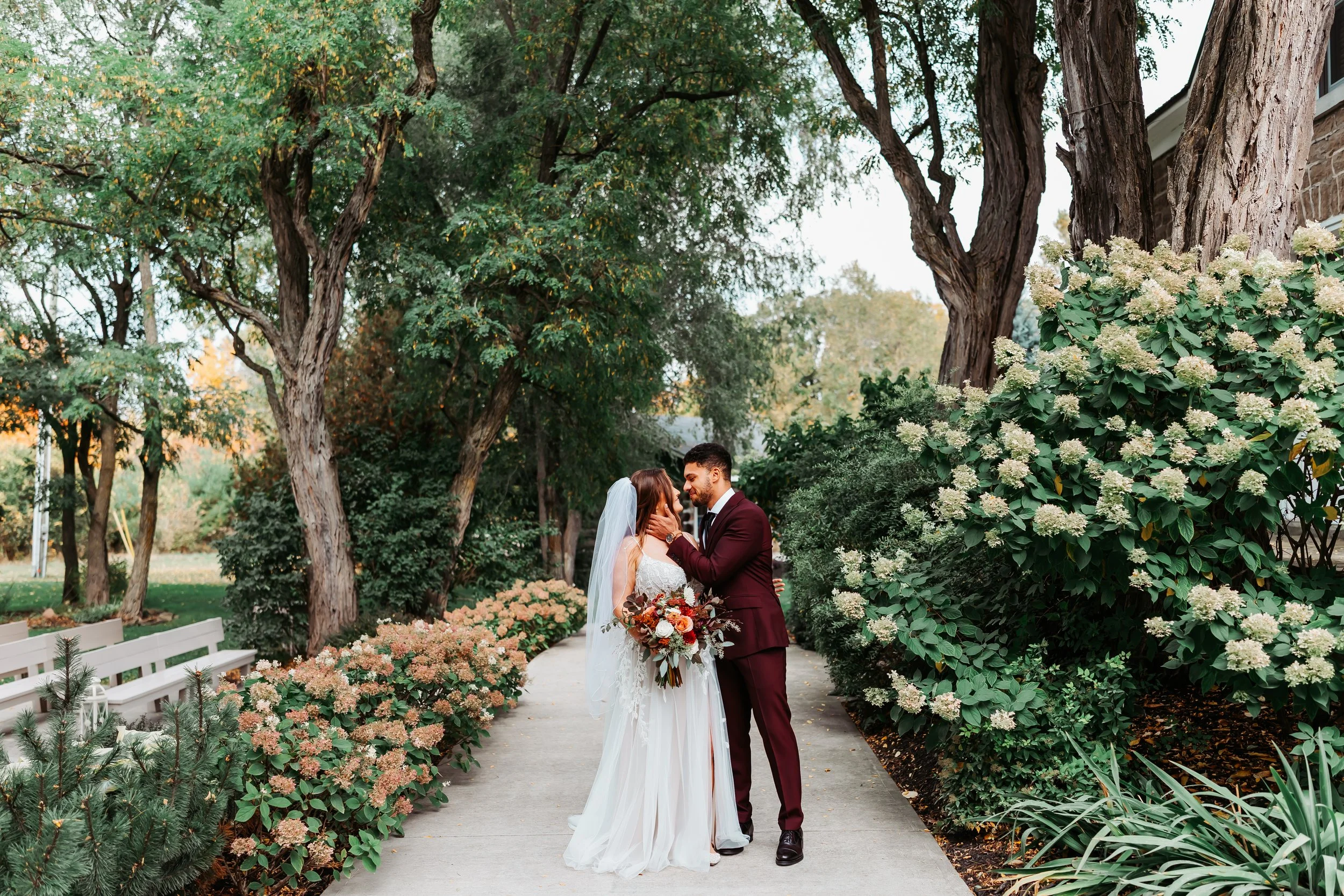 A bride and groom standing on a pathway surrounded by trees and bushes, sharing an intimate moment during their wedding.