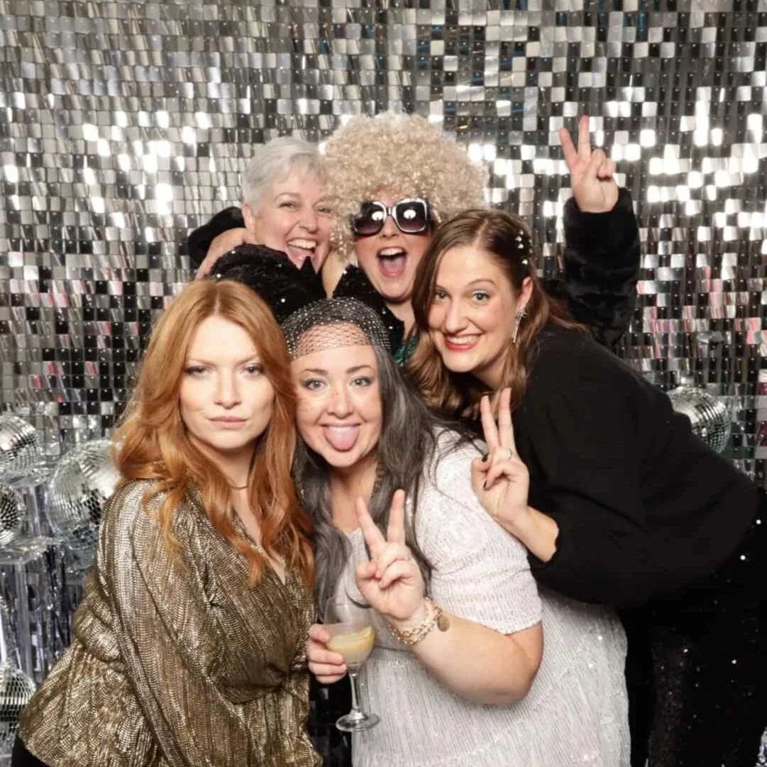 A group of five women celebrating, posing for a photo with a shiny, reflective backdrop and disco balls. They are smiling, making peace signs, and wearing festive clothing.