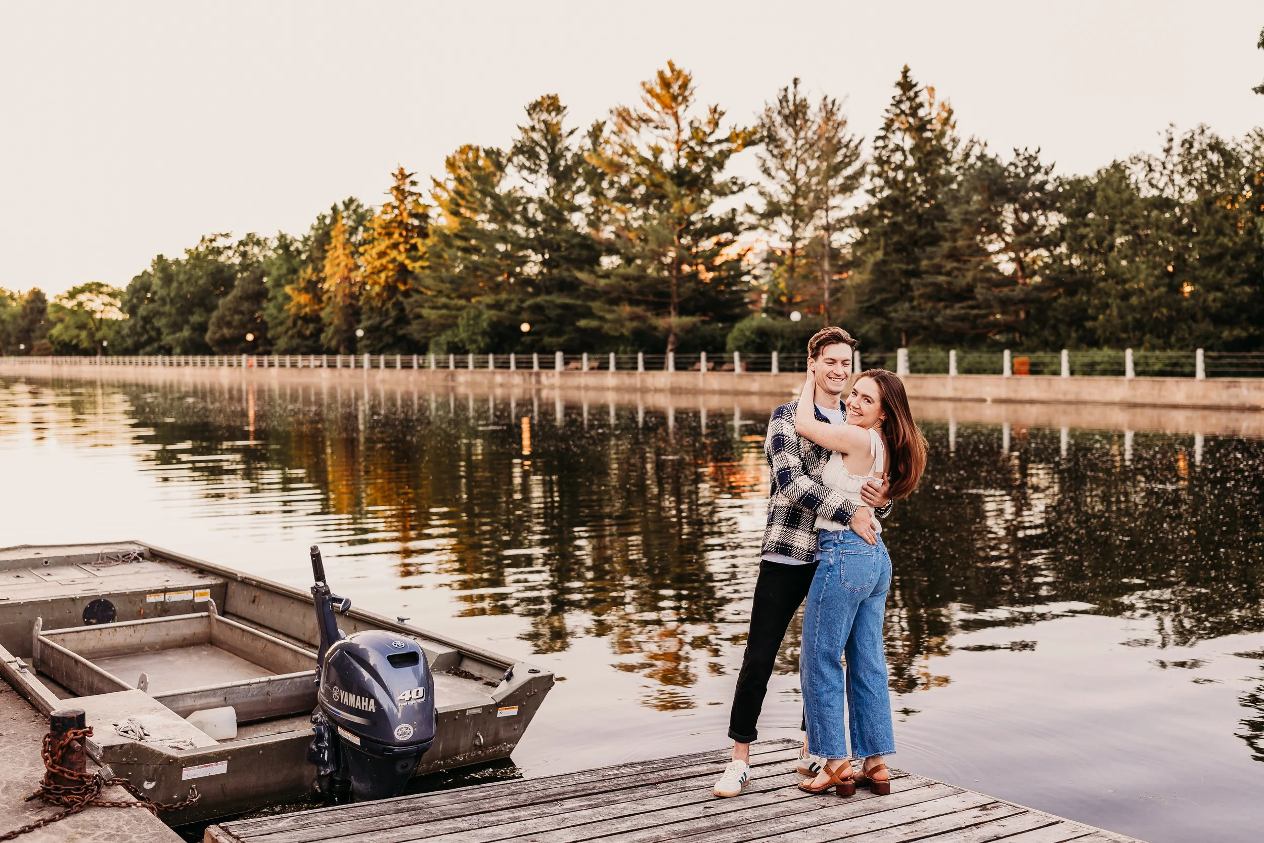 A happy couple embracing on a wooden dock by a lake during sunset, with trees and a boat with an outboard motor in the foreground.