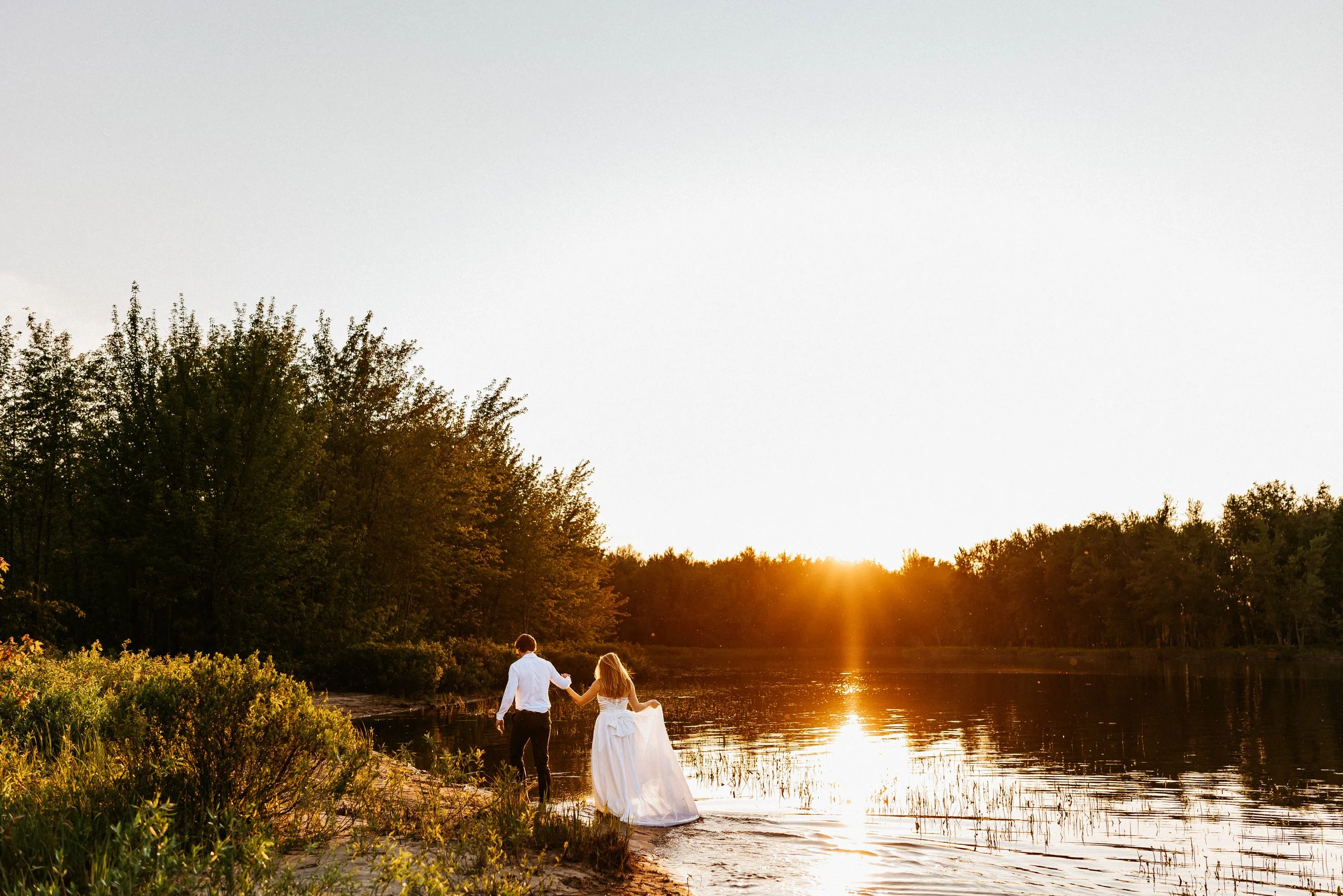 A couple walking hand in hand along the shore of a lake during sunset, with the bride in a white wedding dress and the groom in a white shirt and dark pants, surrounded by trees.