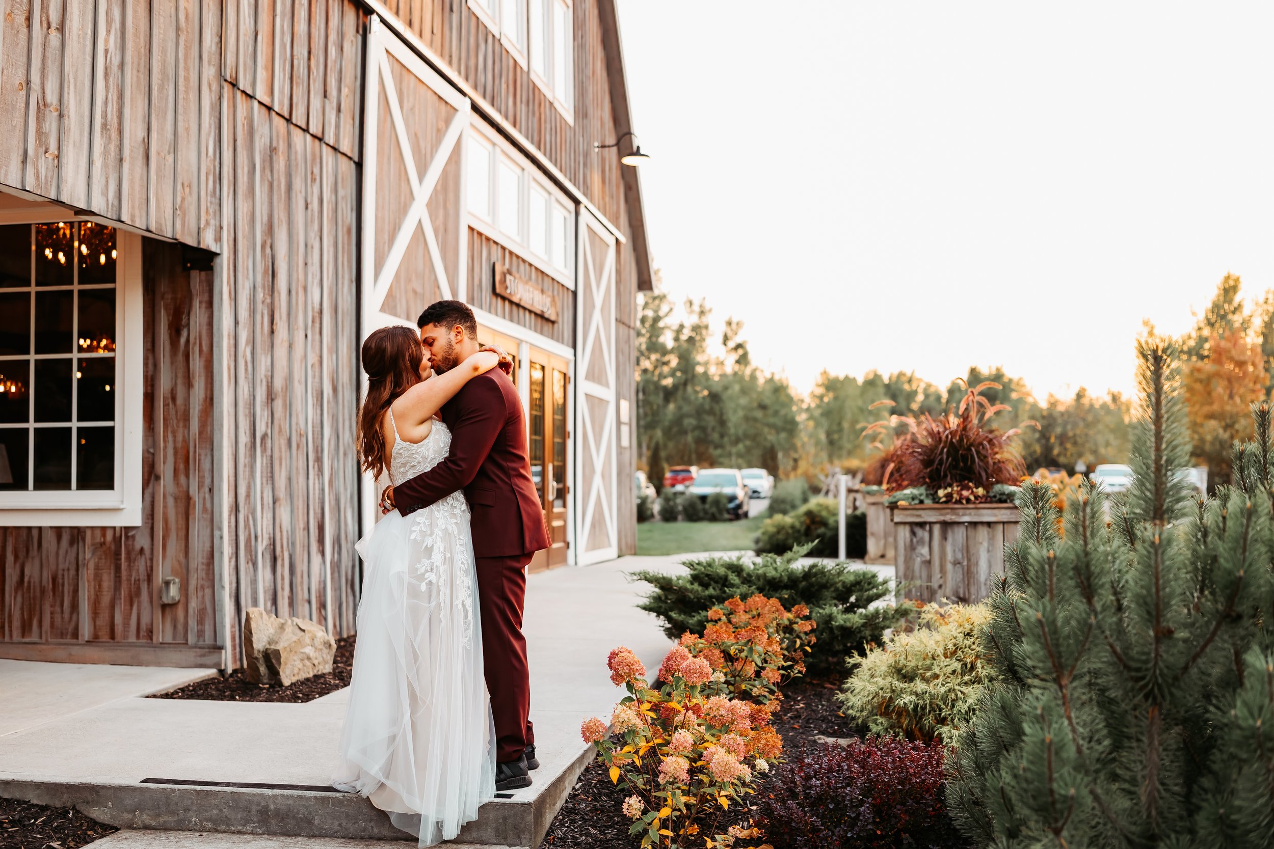 A bride and groom are embracing and kissing outside a rustic wooden building during sunset, with trees and flowers around them.
