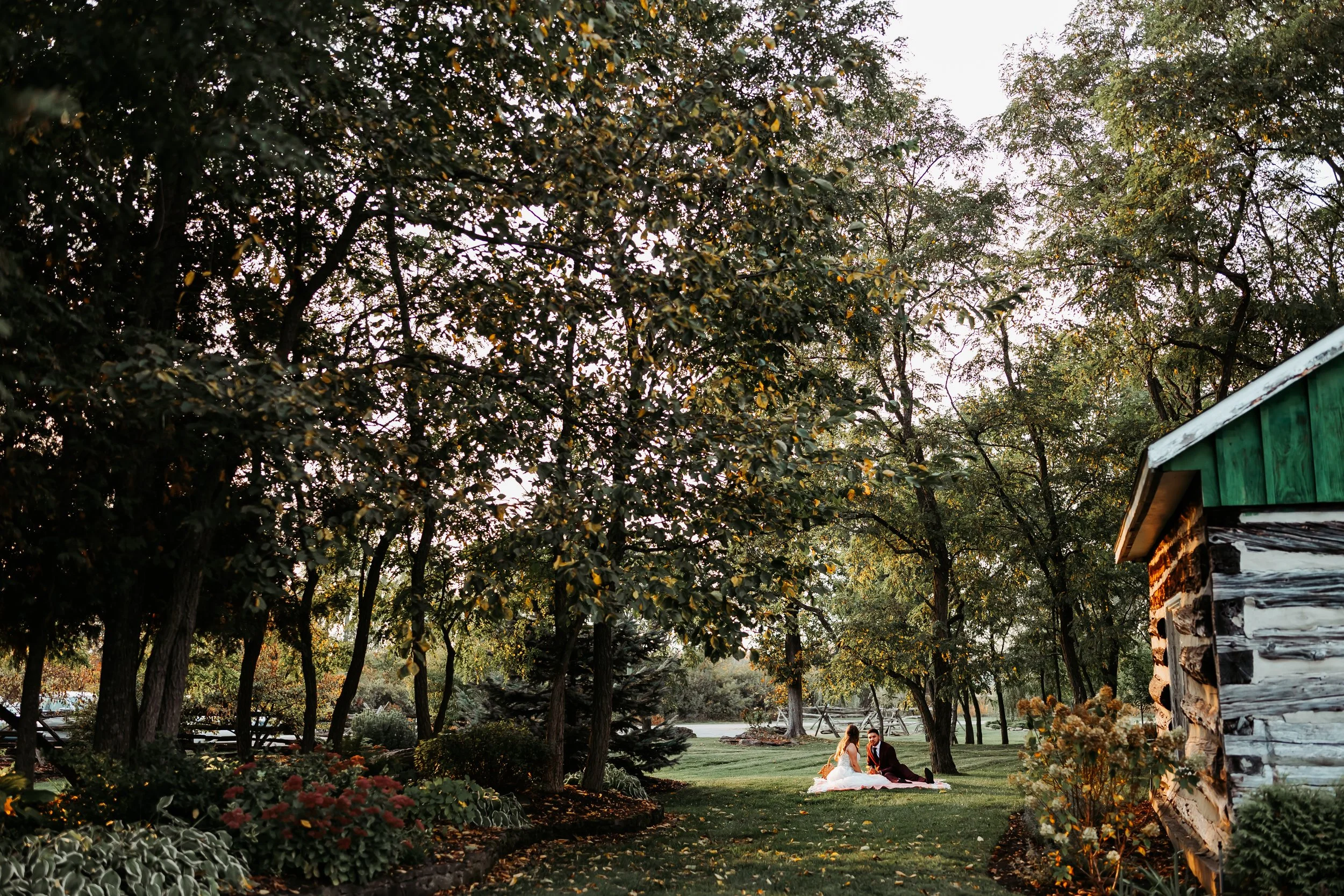 Bride and groom sitting on a blanket in a lush, green park surrounded by trees and flowers, during sunset.