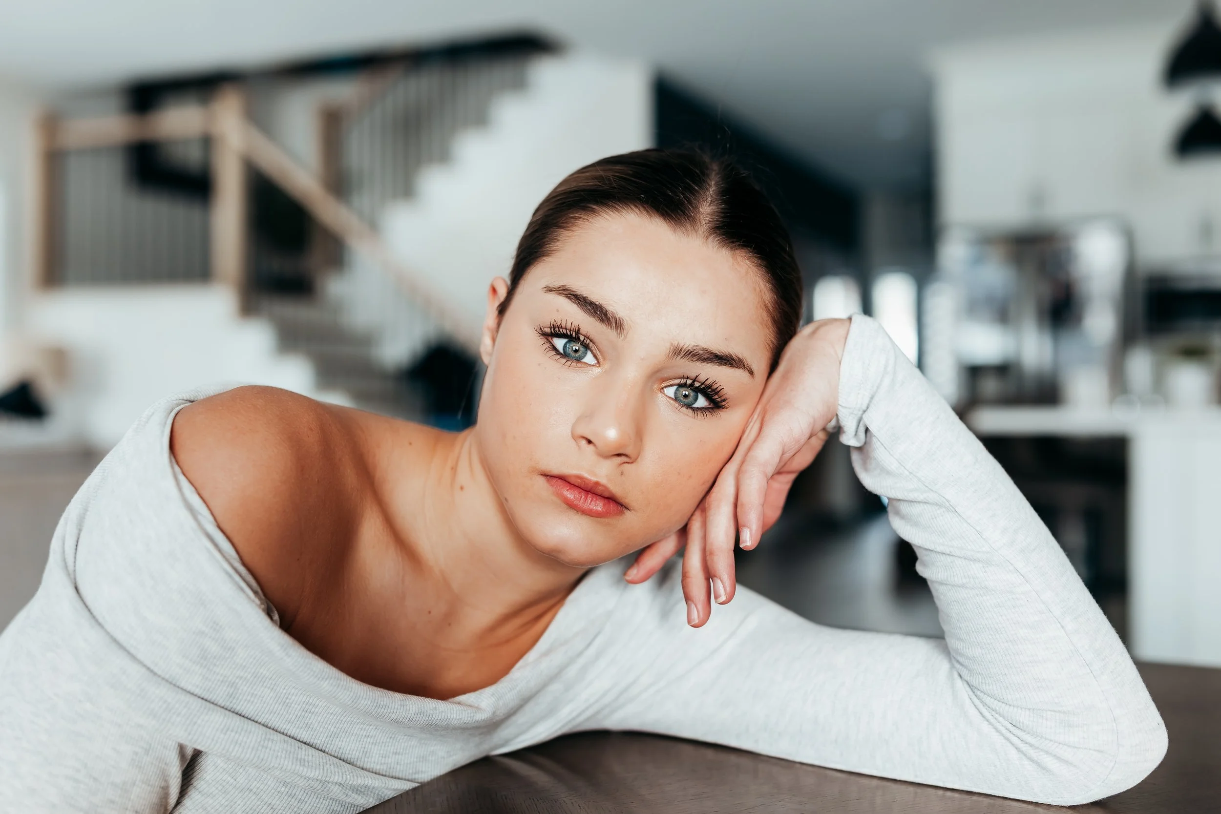 A woman with blue eyes and dark hair resting her head on her hand at a table in a modern home.