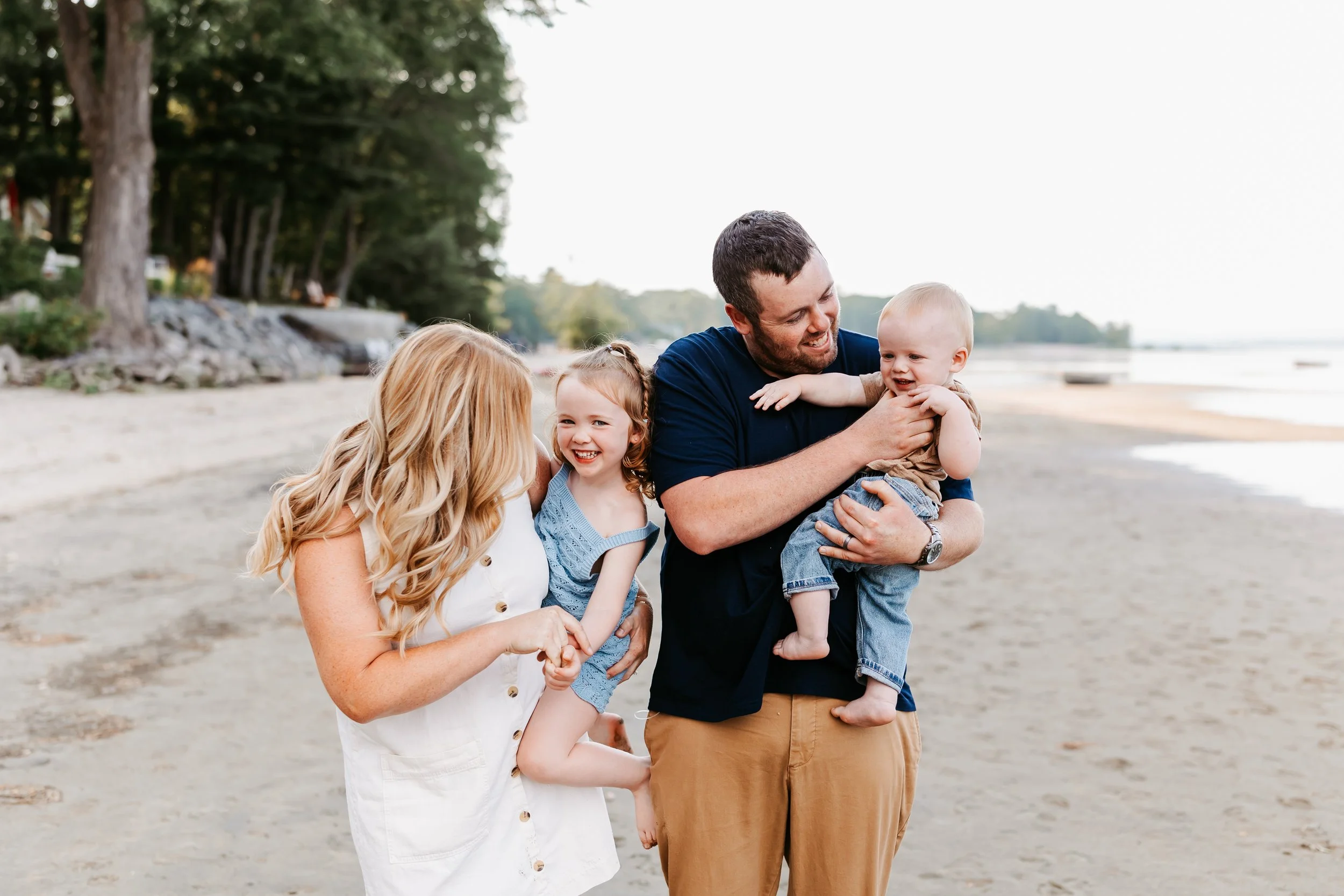 A family enjoying a day at the beach, with a woman, two young girls, and a man playing and laughing on the sand near the water, surrounded by trees.