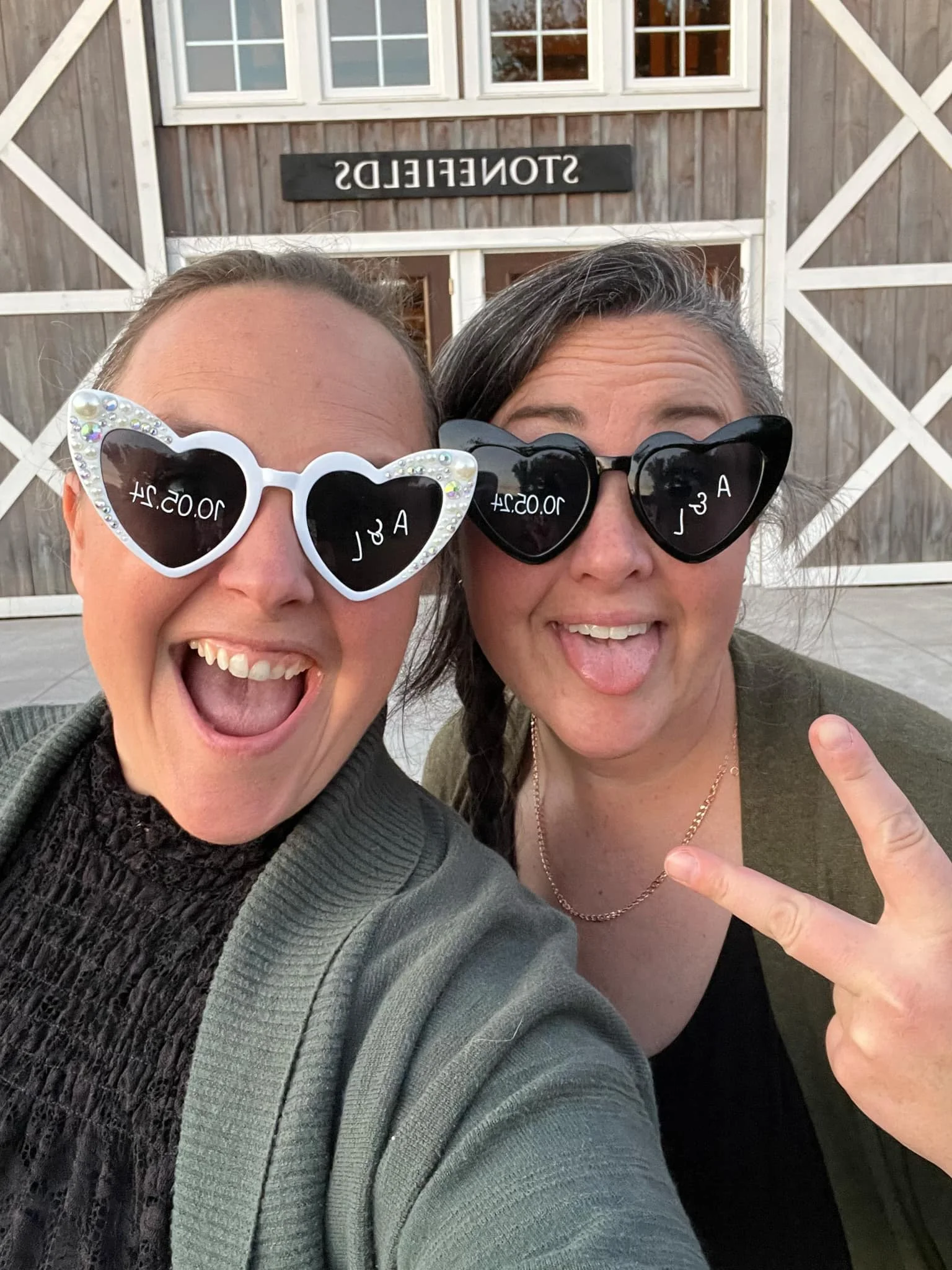 Two women wearing heart-shaped sunglasses, smiling and making peace signs in front of a building with a sign that reads "STONEFIELDS."
