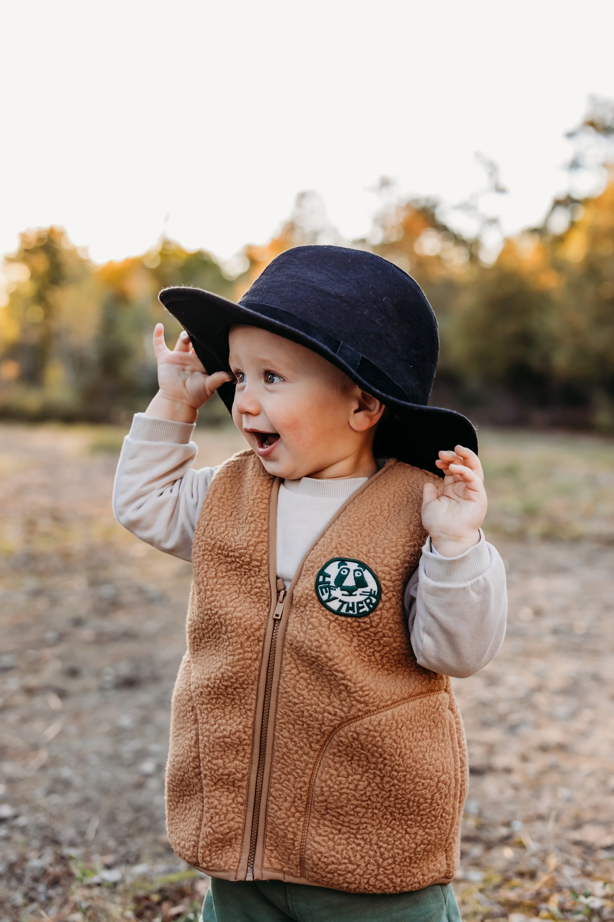 A young boy wearing a black wide-brimmed hat, a beige long-sleeve shirt, and a brown fleece vest with a patch, standing outdoors on a dirt path with autumn trees in the background.