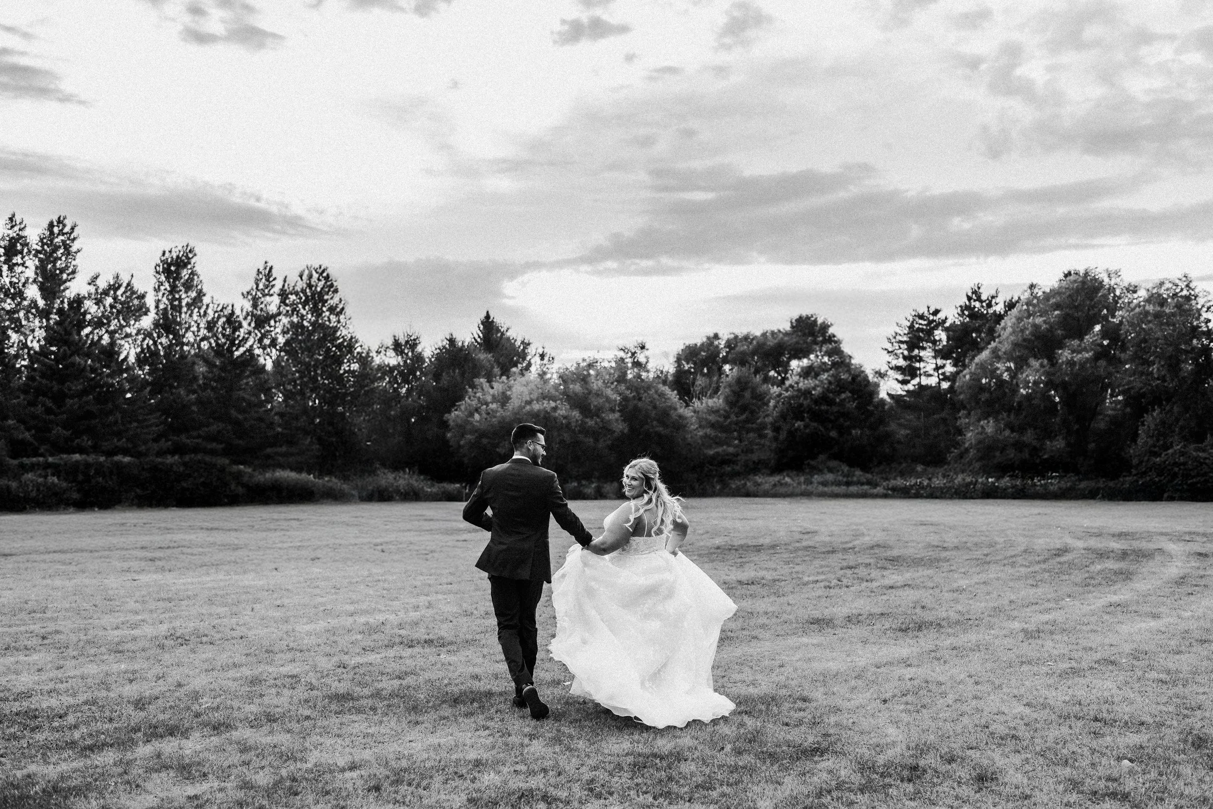 A black and white photo of a bride and groom holding hands and walking across a grassy field with trees in the background, during sunset.