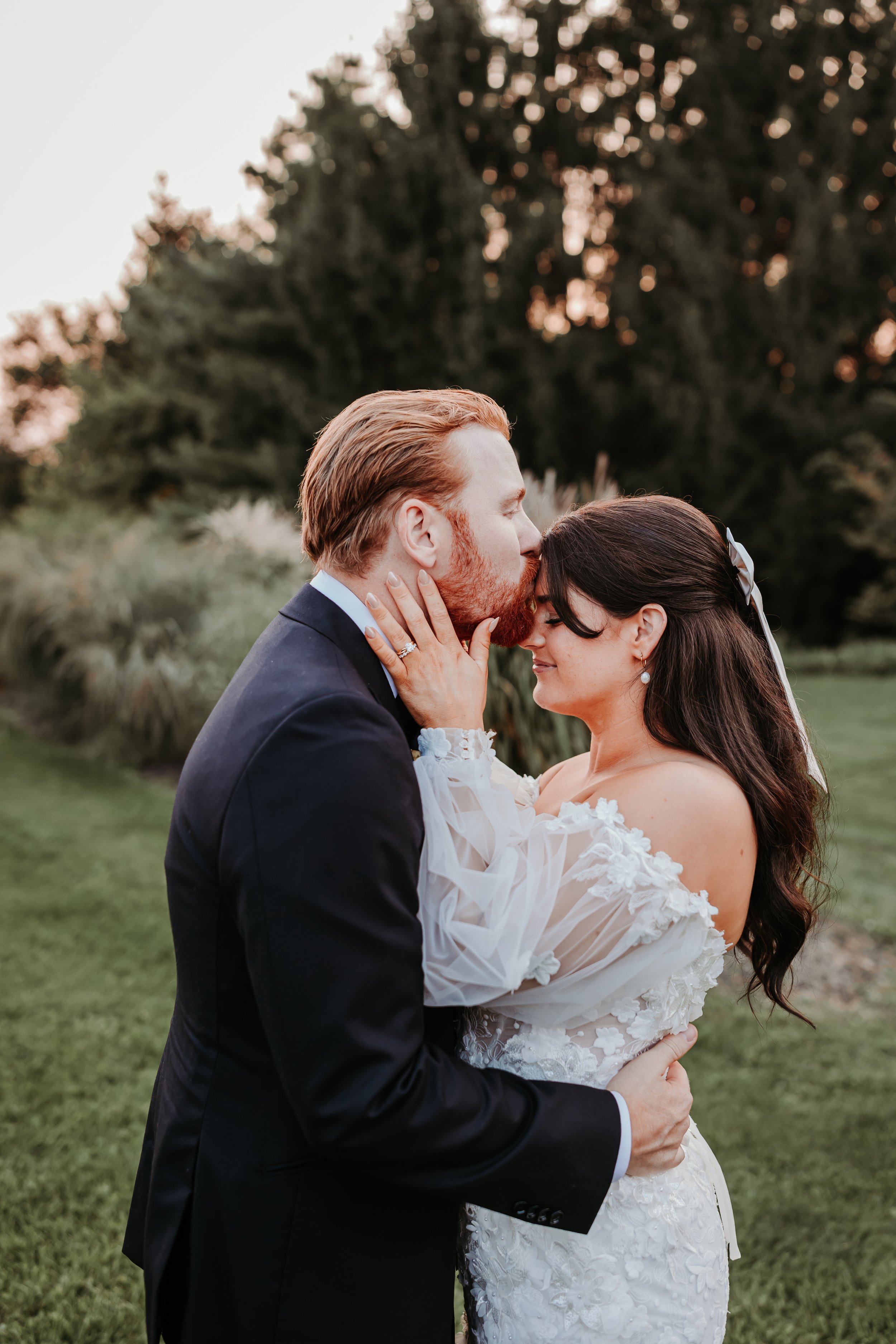 A bride and groom share an intimate moment outdoors, with the groom kissing the bride's forehead. The bride has dark hair and is wearing a white lace wedding dress, while the groom has red hair and a beard, dressed in a dark suit. The background features greenery and trees during sunset.