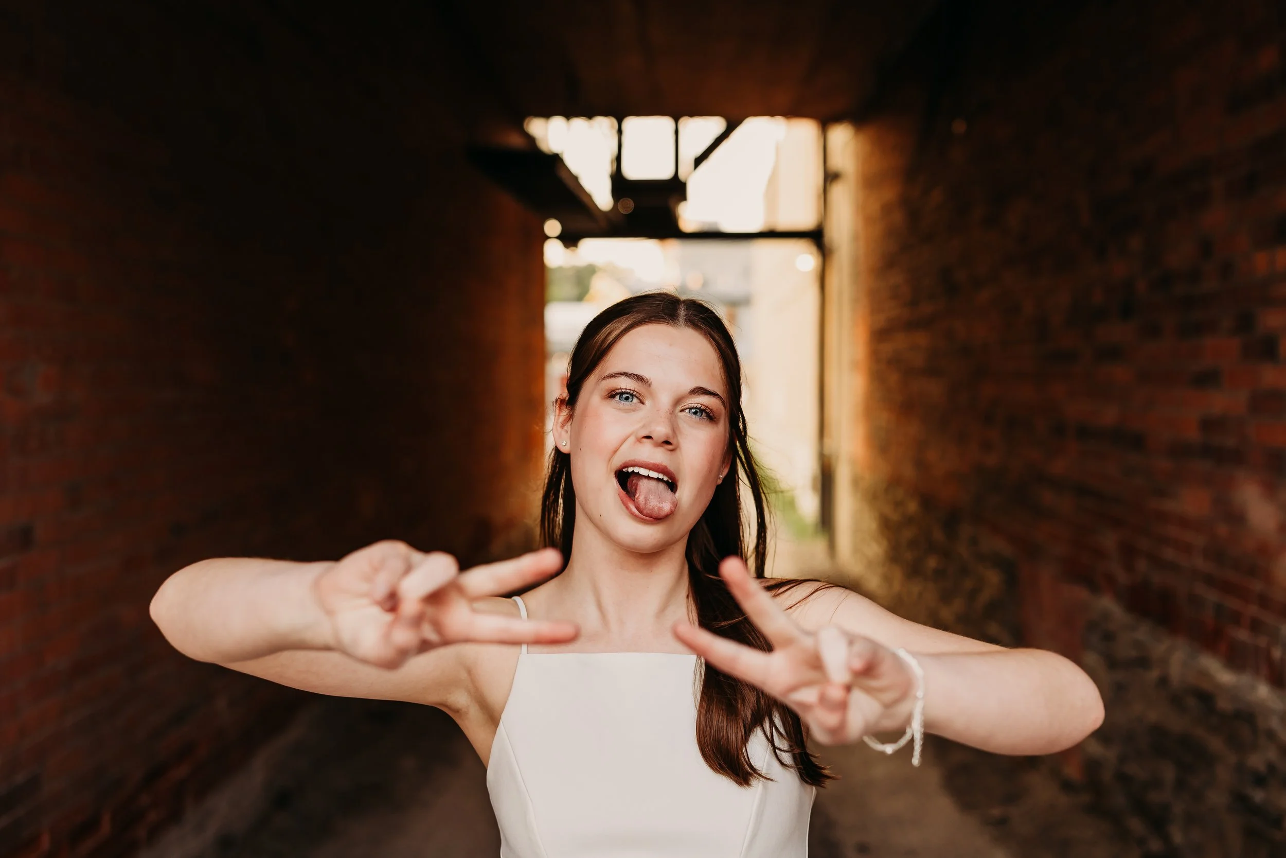 Young woman standing in a narrow alleyway, making peace sign gestures with both hands, sticking out her tongue, and smiling with her mouth open. She has brown hair, blue eyes, and is wearing a white sleeveless top.