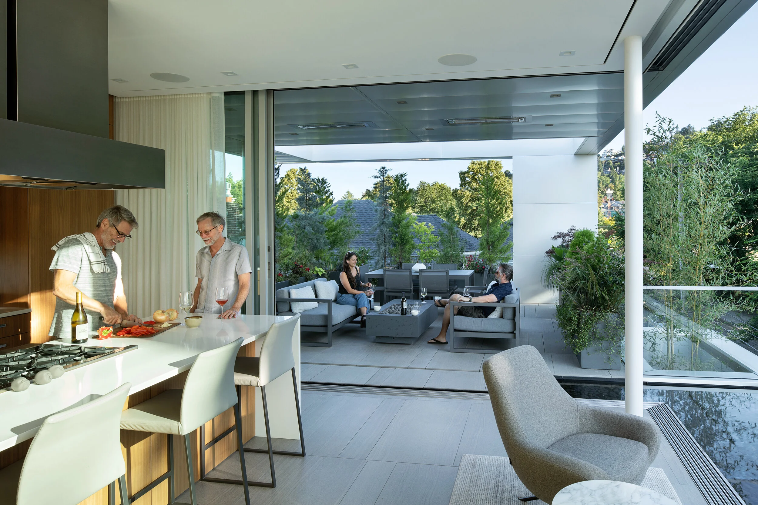 Two men cooking in modern kitchen open to terrace with two people sitting around firepit — William Kaven Architecture