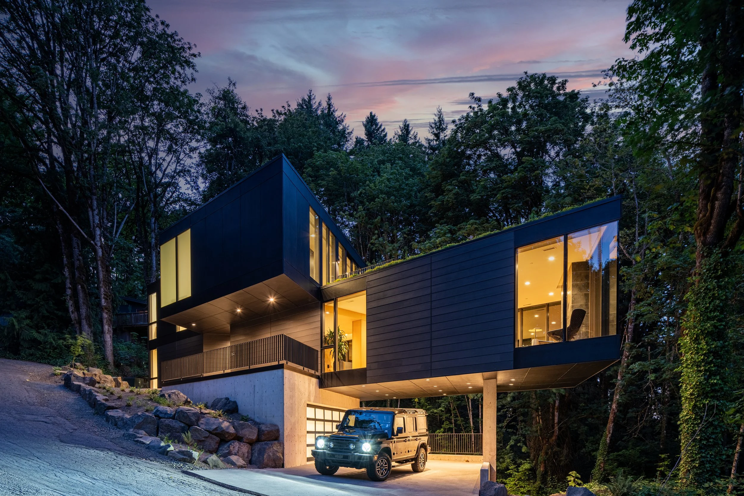 Modern house in forest at dusk with carport and cantilevered green roof — William Kaven Architecture