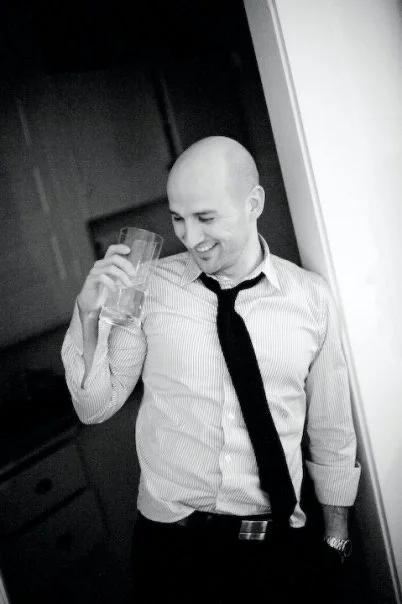 A bald man in a striped dress shirt and black tie holding a glass of water, smiling and looking down.