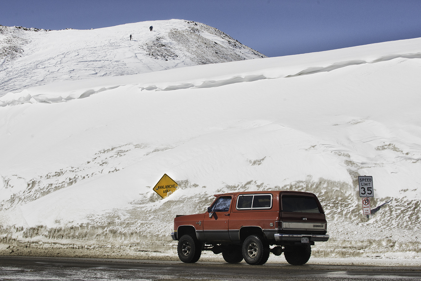 A Chevy Blazer crosses over the top of the pass in front of a buried Avalanche Area sign. Snowboarders can be seen up on the hill, hiking for a good spot to drop in.