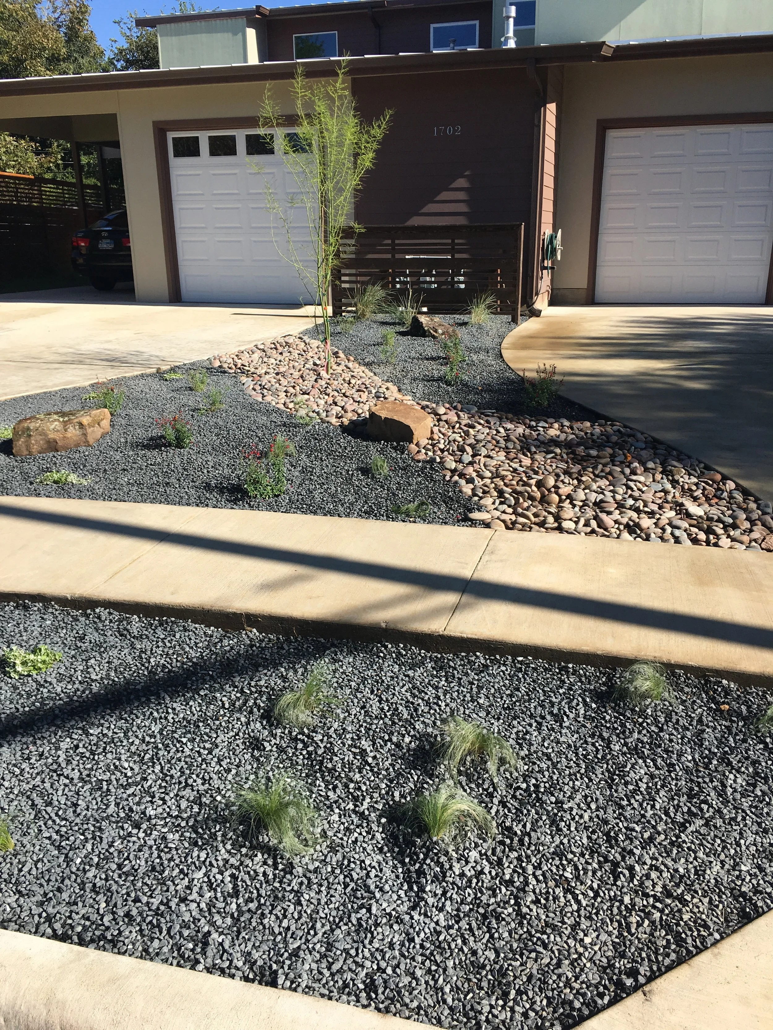 Modern front yard with xeriscaping, featuring gravel, stones, small plants, and a tree, adjacent to a driveway and garage.