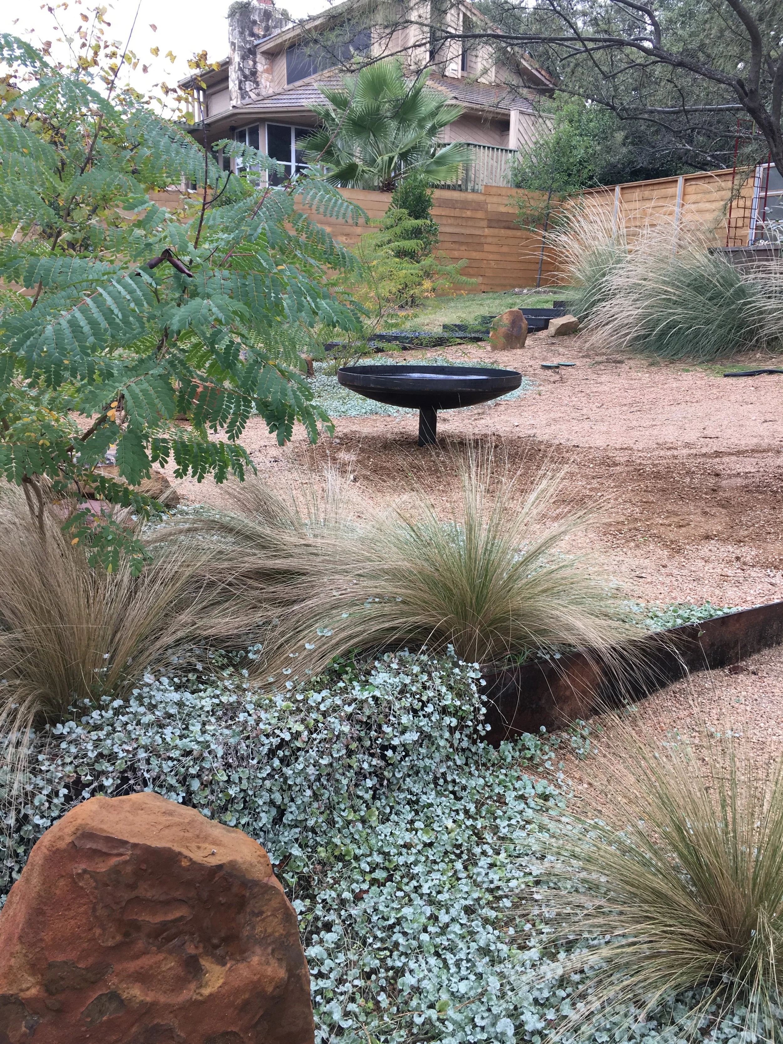A landscaped garden with a variety of plants, including grasses and leafy shrubs. A round black metal fire pit is situated in the center. A house with a stone chimney is visible in the background, surrounded by a wooden fence and tall trees.
