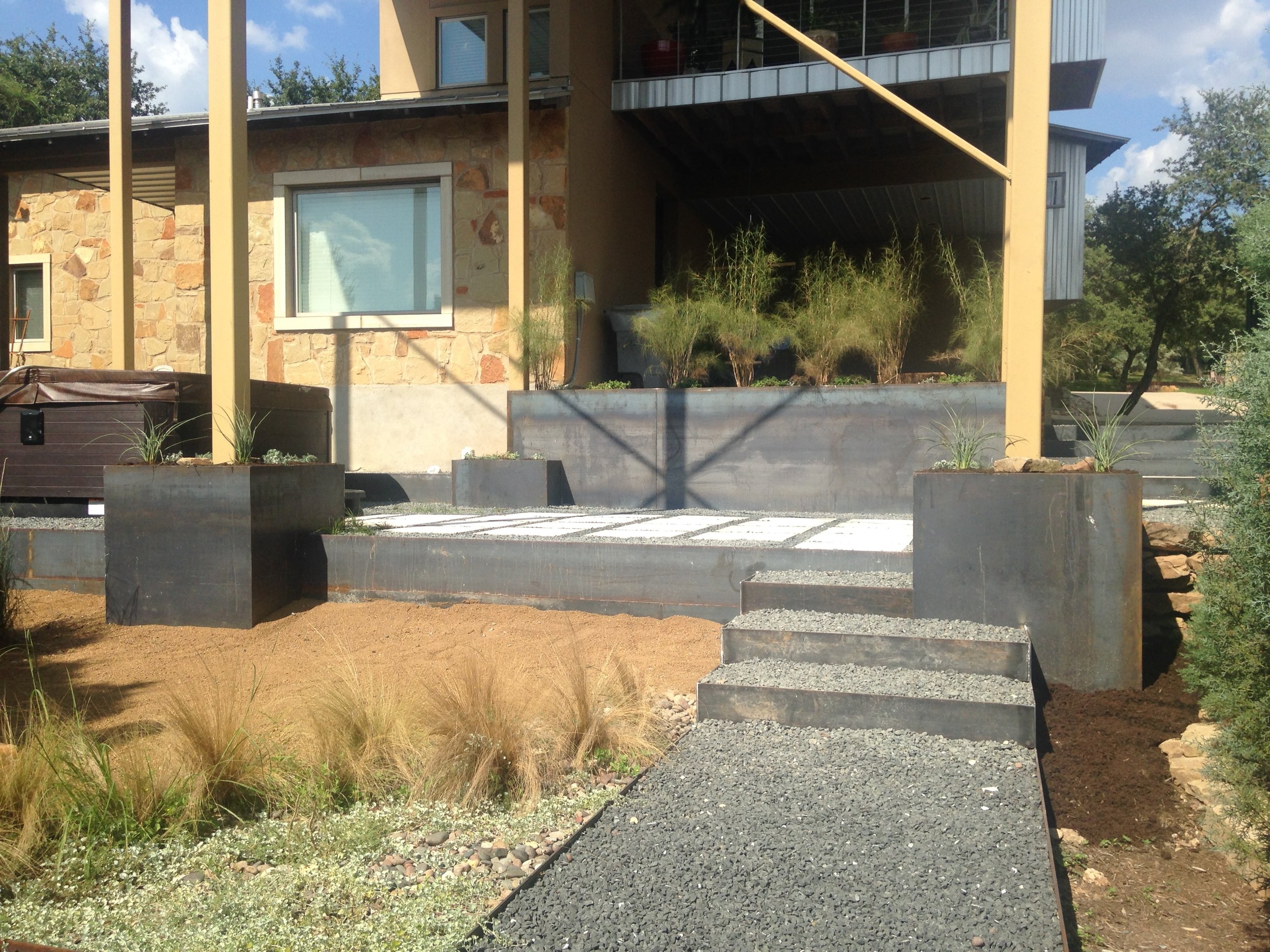 Modern outdoor patio design with raised planters and gravel walkway in front of a stone-clad building.