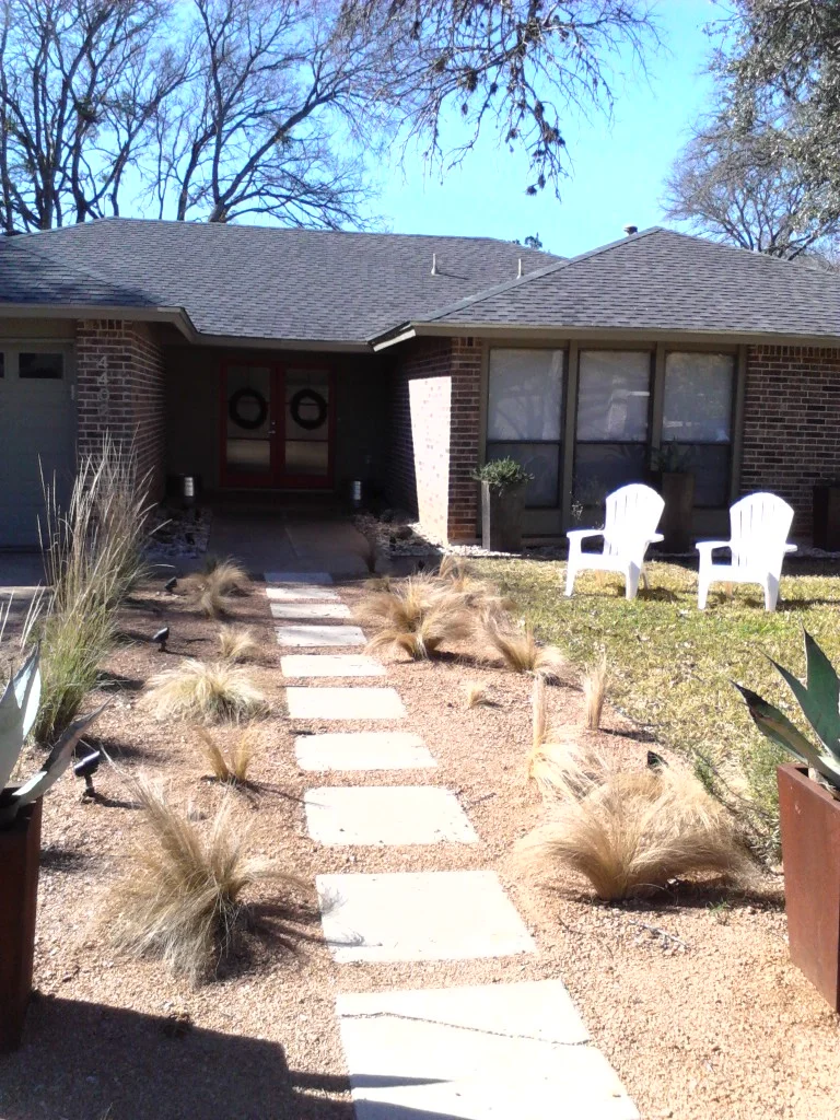 Pathway leading to a brick house with red double doors, surrounded by desert landscaping and two white chairs.