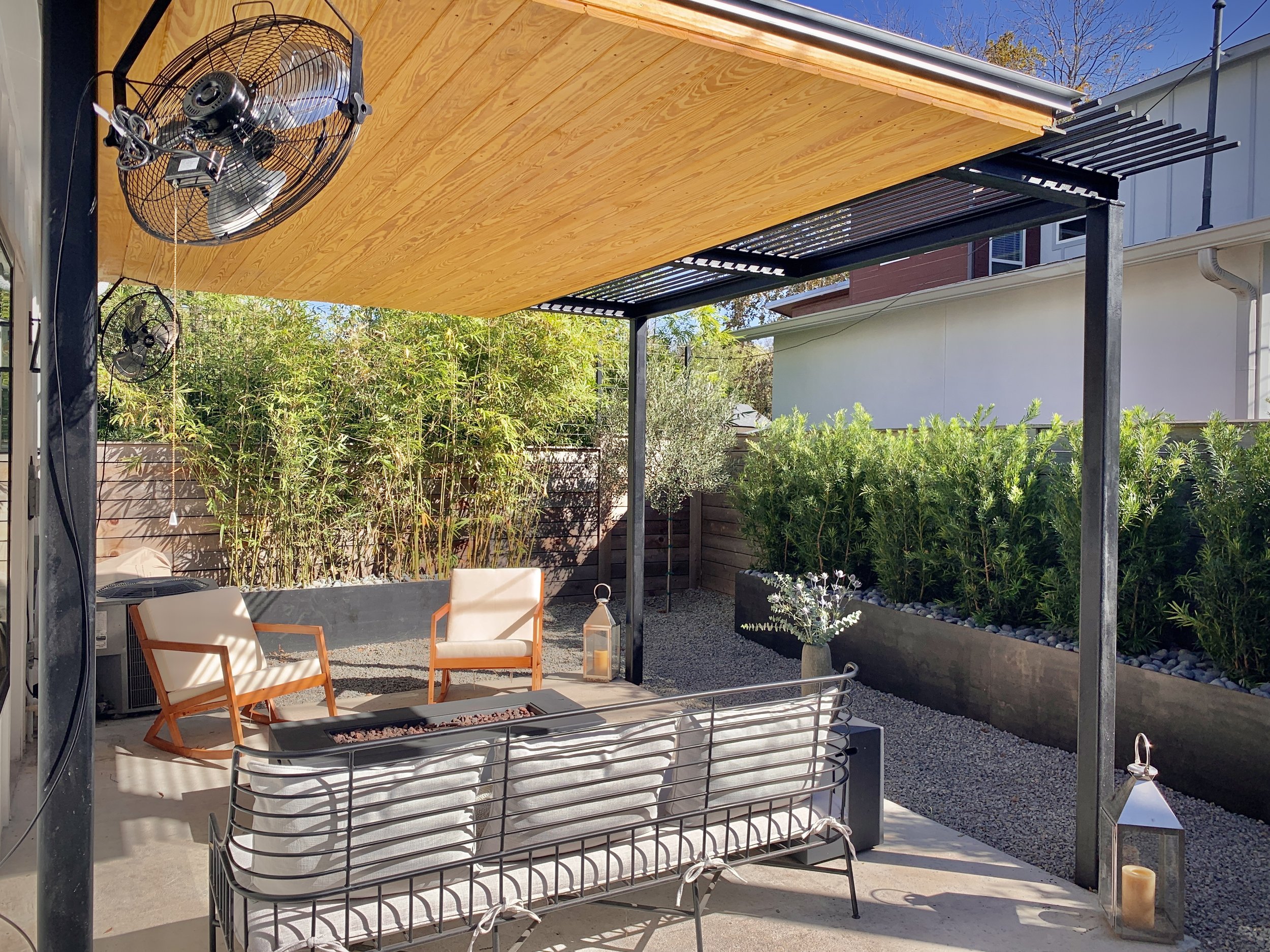 Outdoor patio area with wooden pergola, ceiling fans, two chairs, a metal bench, a fire pit table, lanterns, and surrounding greenery.