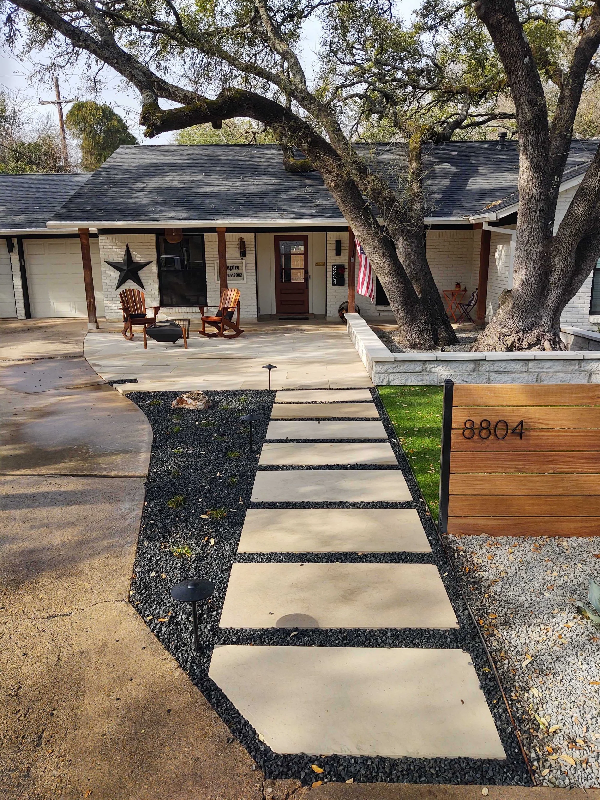 Modern house exterior with stone walkway, two large trees, wooden chairs on the porch, a star wall decoration, and an American flag. Address number 8804 on wooden panel.