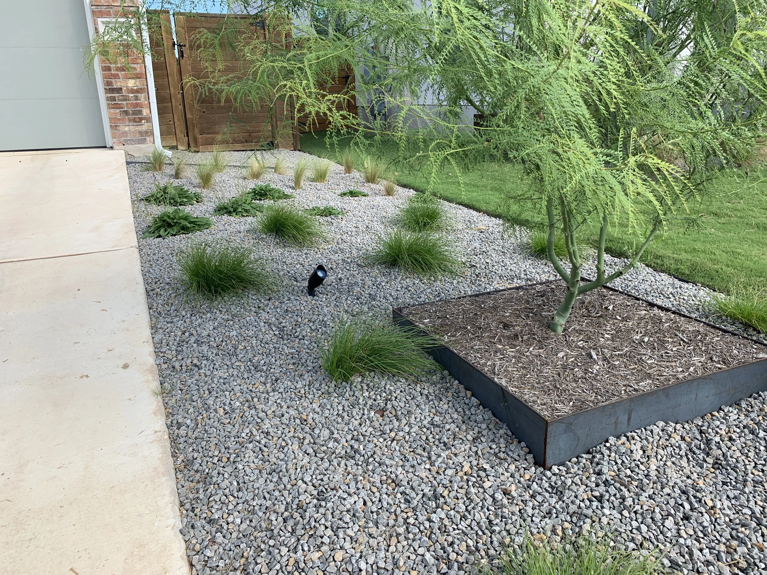 Front yard with xeriscaping, featuring gravel, small plants, a young tree with mulch around its base, and a solar garden light. A wooden fence is in the background.