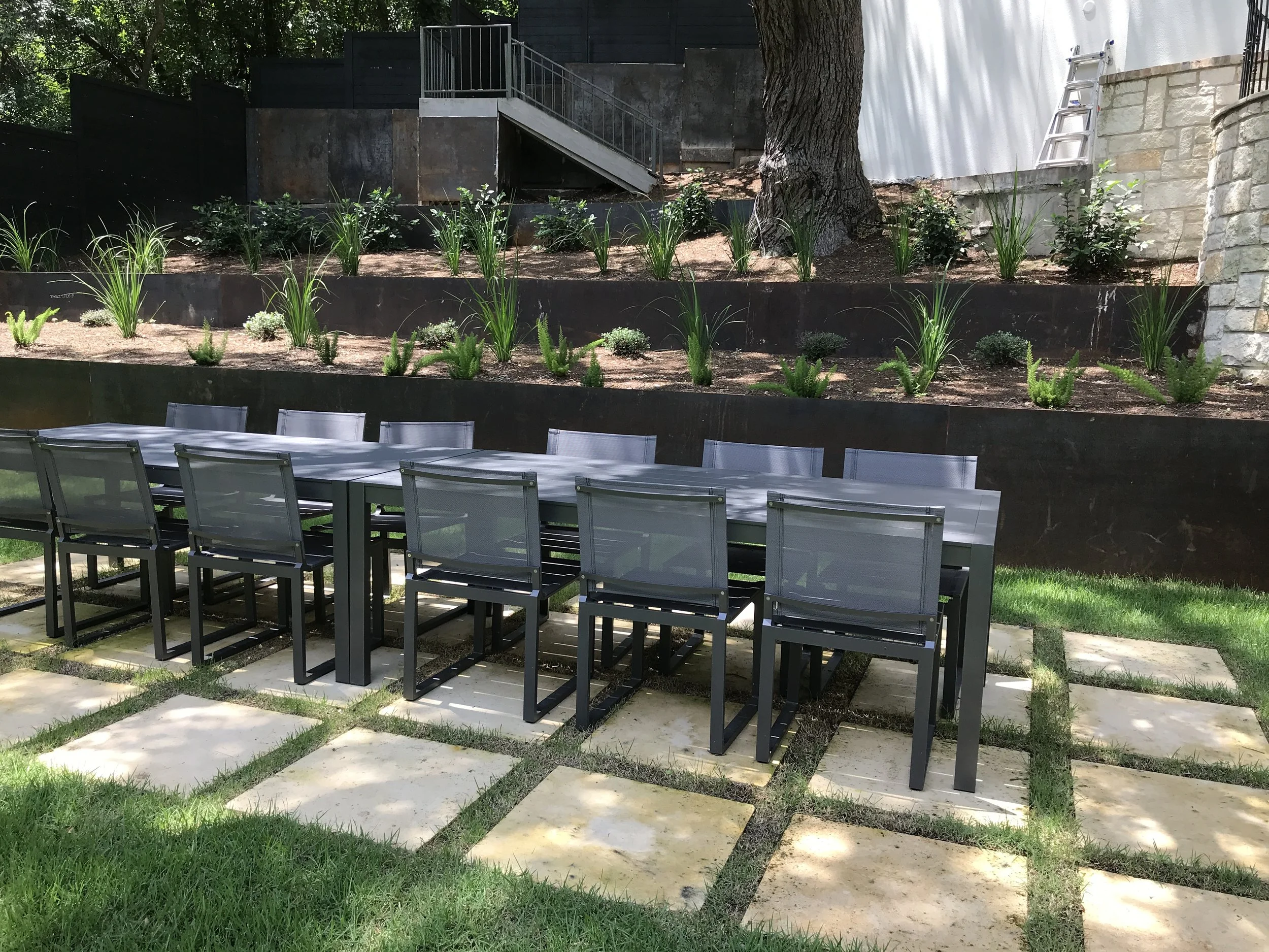 Outdoor dining area with a long table and chairs on a patio, surrounded by greenery and landscaped retaining walls.