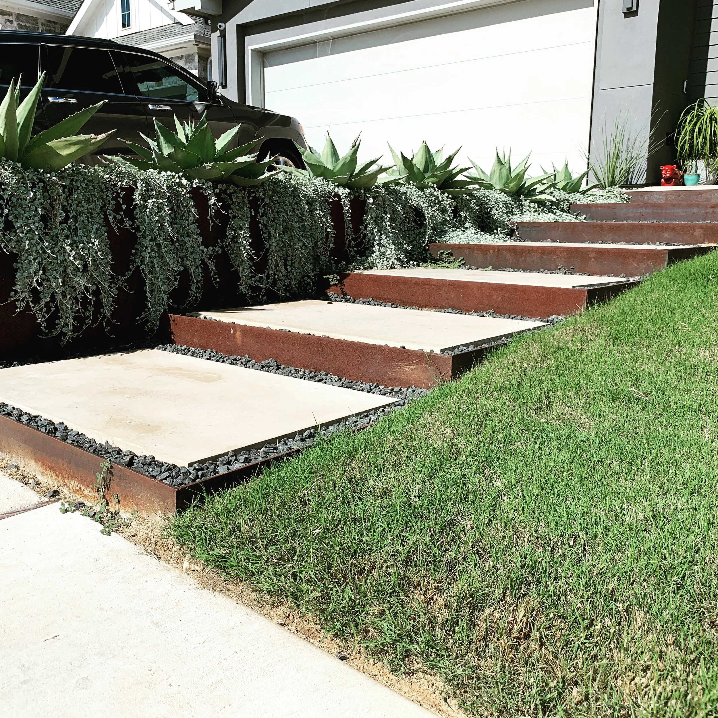 Modern outdoor staircase with concrete steps, surrounded by greenery and succulents, next to a driveway.
