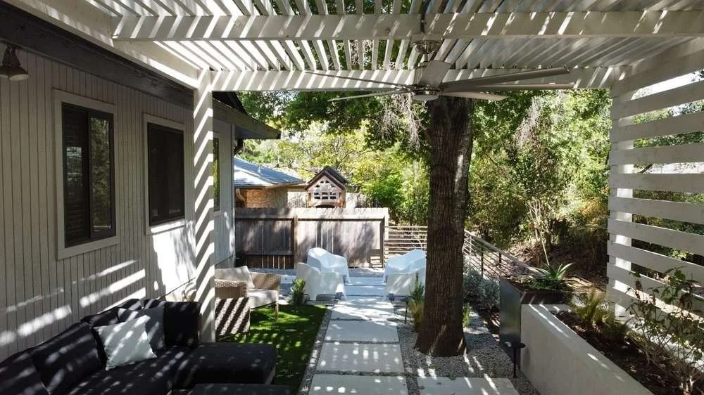 Outdoor patio area with a shaded pergola, black sofa with pillows, white chairs, a tree, and garden plants, adjacent to a house with beige siding.