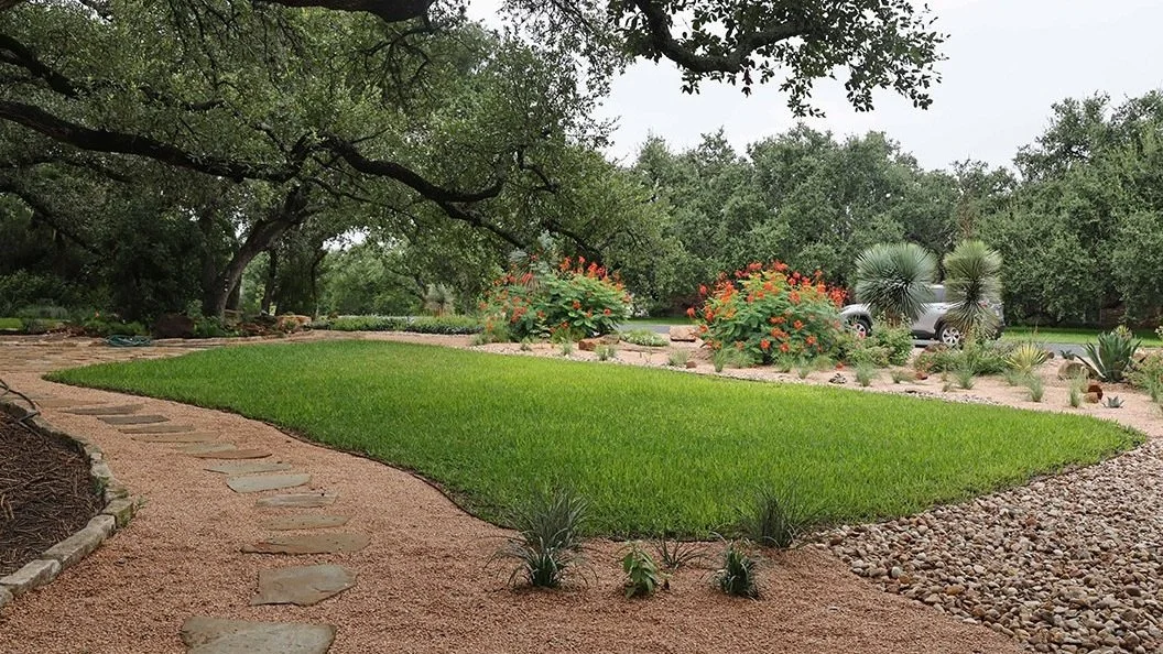 A landscaped garden with a green grass patch, stone pathway, decorative bushes, flowering plants, and trees under an overcast sky.