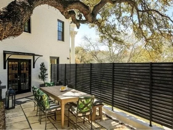Outdoor patio area with a wooden dining table surrounded by green and beige chairs. A black privacy fence encloses the space, with a large tree providing shade. A white building with windows and a door is visible on the left.