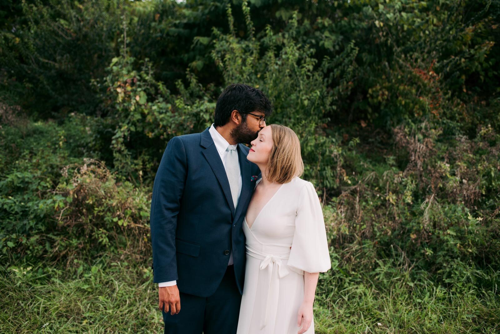Groom kissing bride's forehead before lush greenery in Pittsburgh PA Carly Romeo + Co.