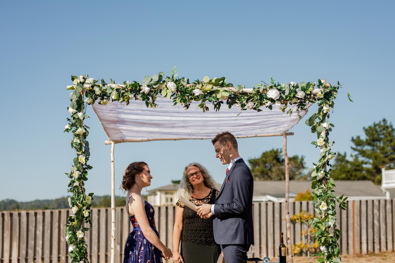 Groom reading vows to bride with officiant under backyard floral chuppah in Mendocino California Carly Romeo