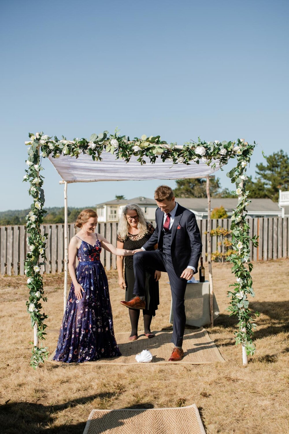 Groom breaking the glass under backyard chuppah with bride and officiant in Mendocino CA Carly Romeo + Co.