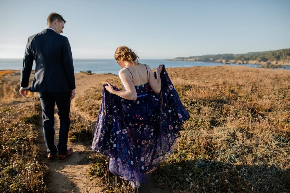 Bride in blue embroidered dress lifting skirt to hike cliff with groom in Mendocino CA Carly Romeo