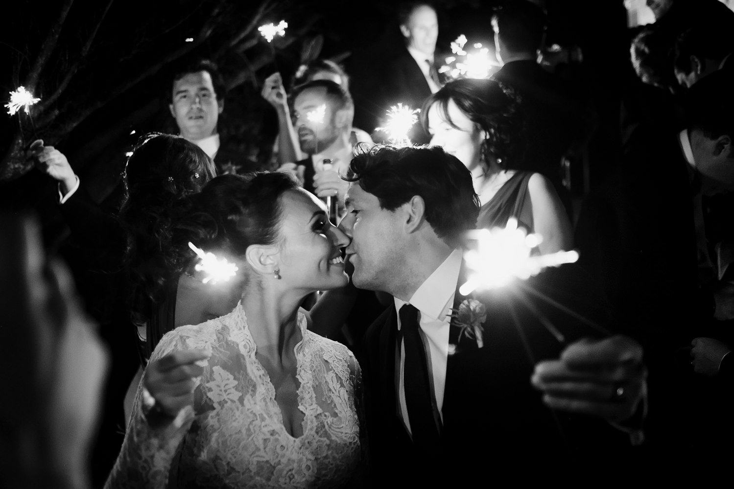 bride and groom kiss as they hold their sparklers away from themselves towards the camera surrounded by their bridal party 