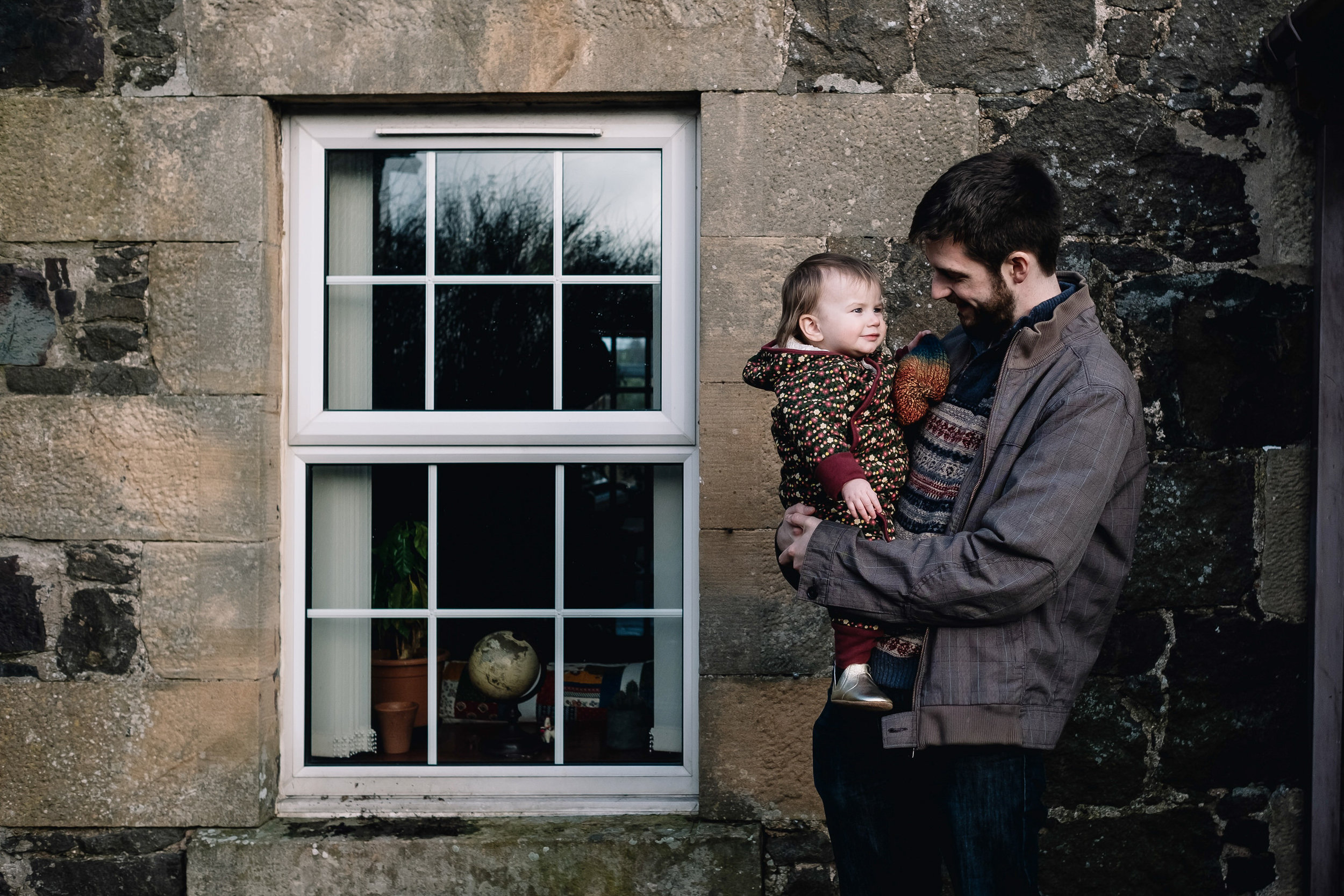 Father and daughter stand outside their house. He is holding her and looking at her.