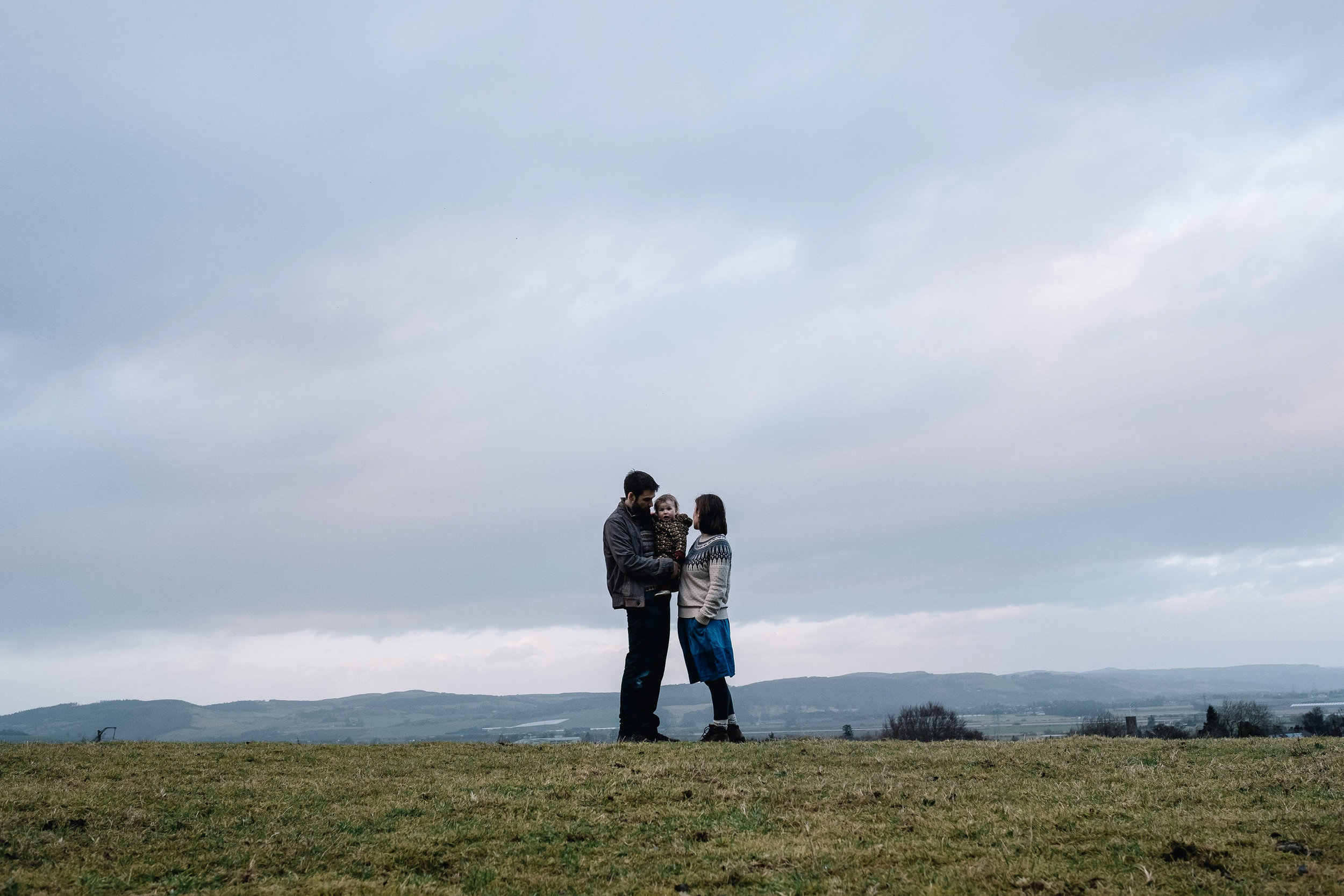 Family of three stand on top of a hill above the city.