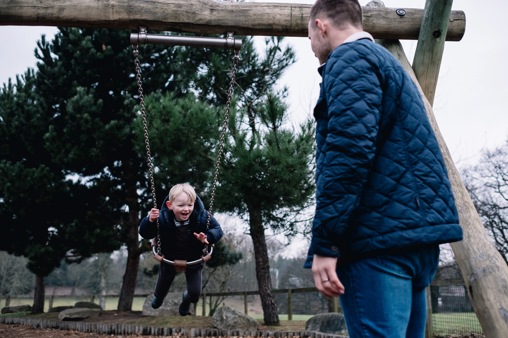 Father stands pushing his son on a swing.