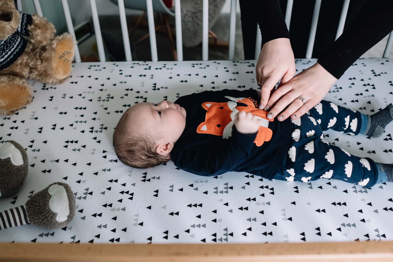 Baby lies in cot and mother's hand are on his stomach.
