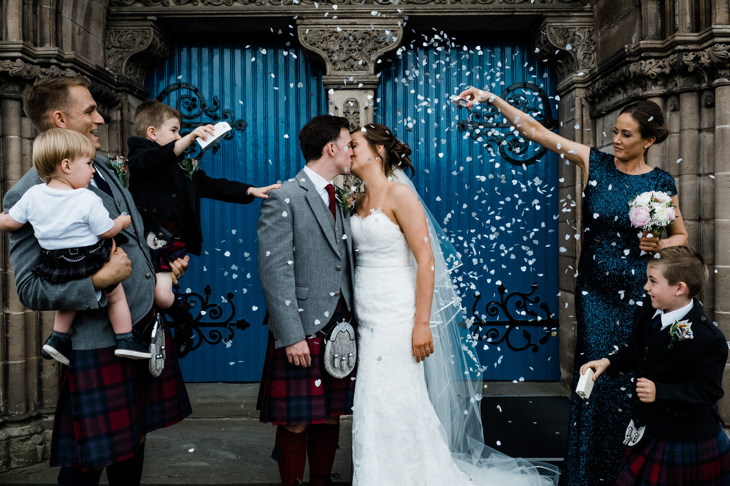  A couple face each other and kiss against a backdrop of two blue doors whilst their closest of kin throw confetti over them. 