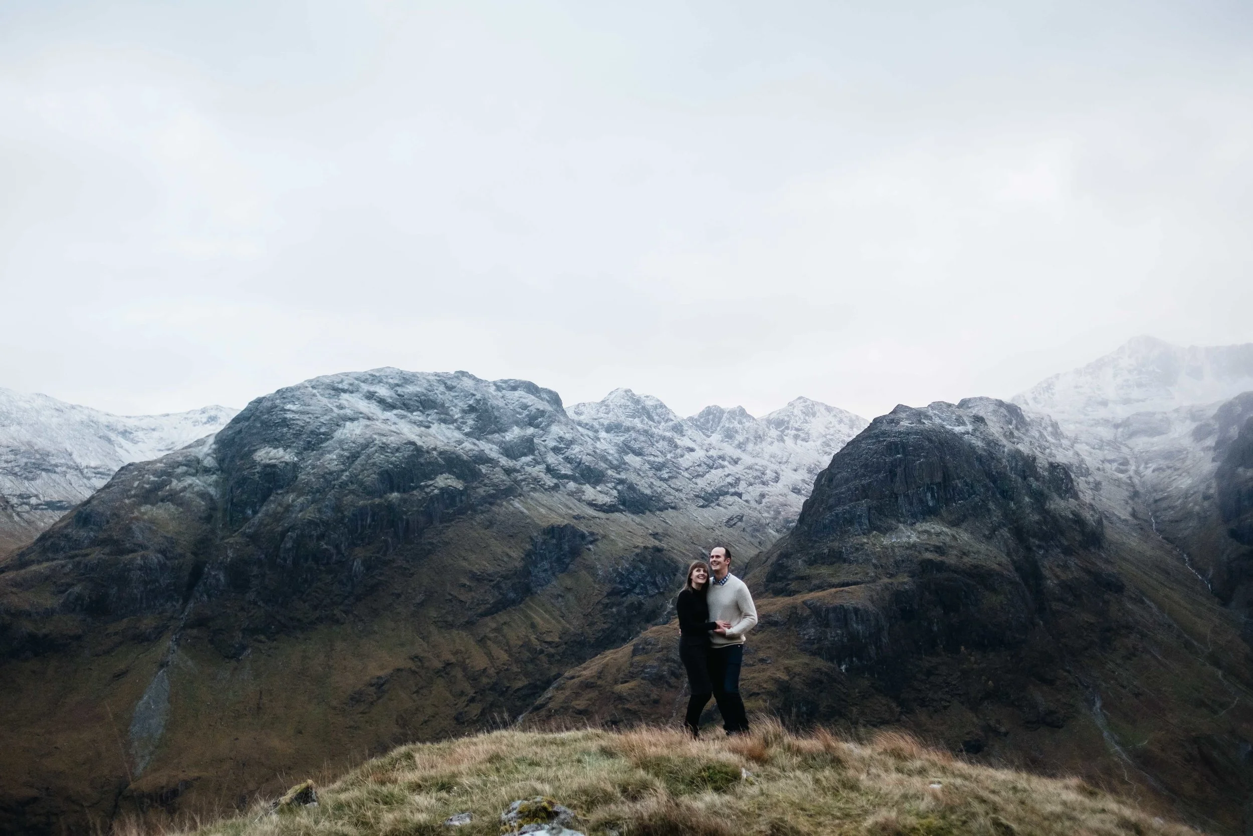 couples standing with snow capped mountains behind in Glencoe