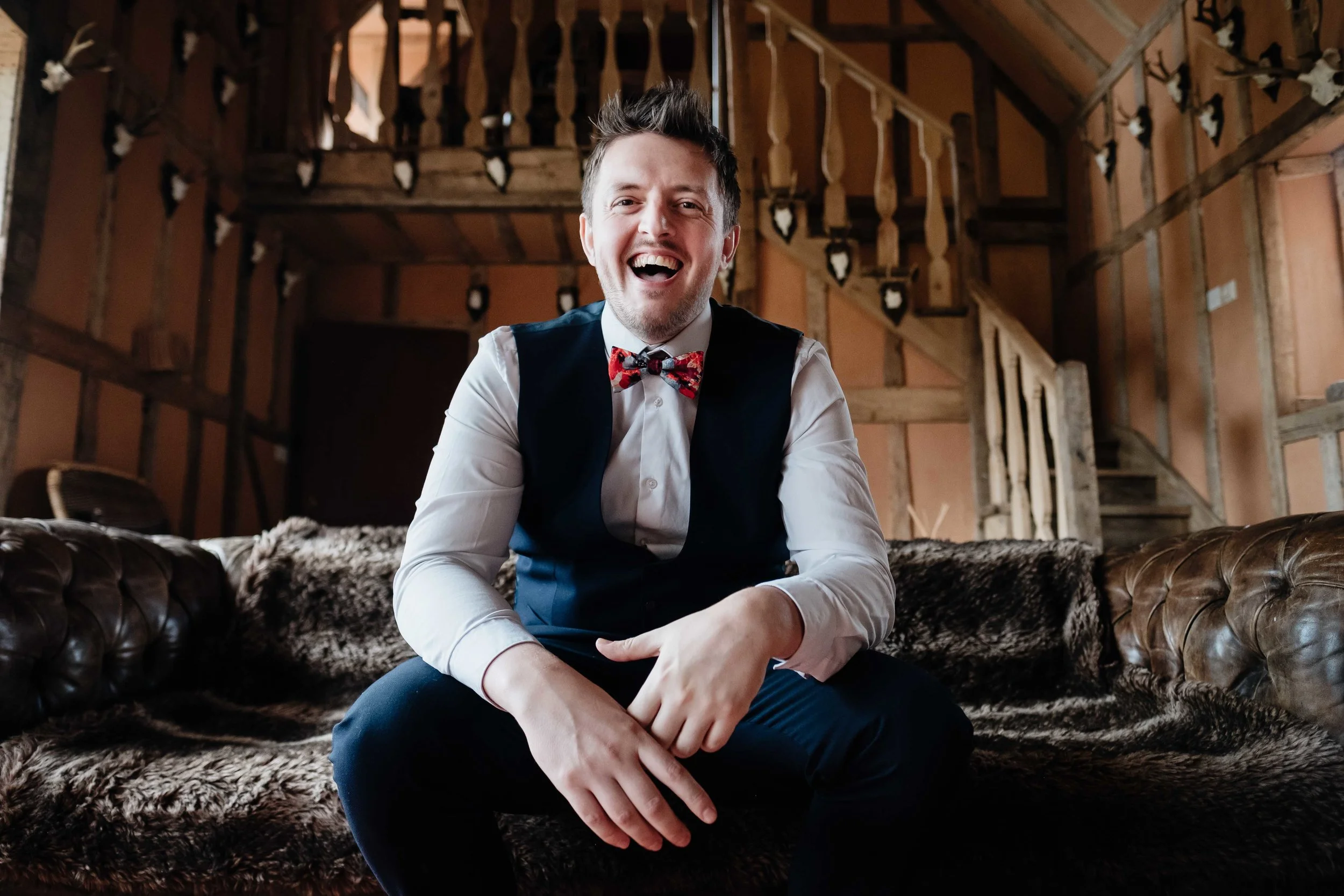 A groom with a suit and bow tie sits laughing on a sofa
