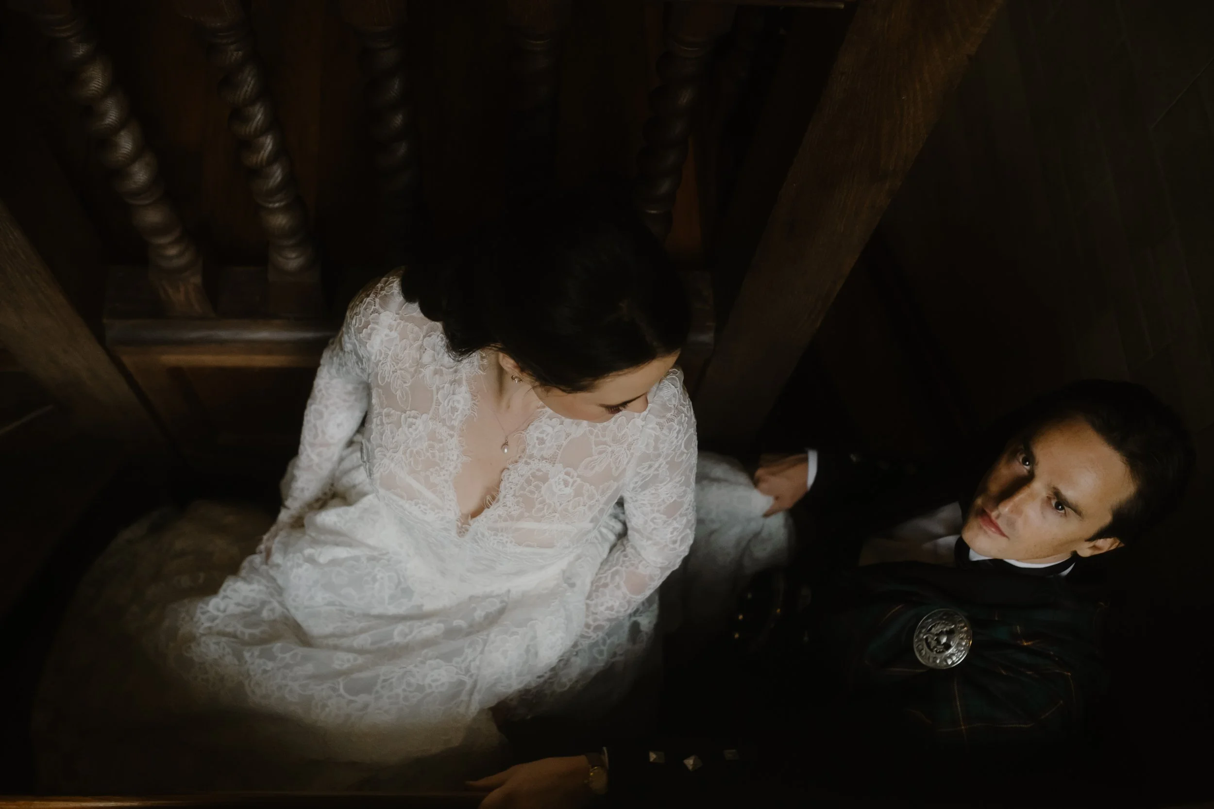 Bride and groom going up a dark gloomy staircase. She is wearing a romantic lace white dress and looking at her husband. He is looking sternly at the camera. Scotland wedding photographer candid.