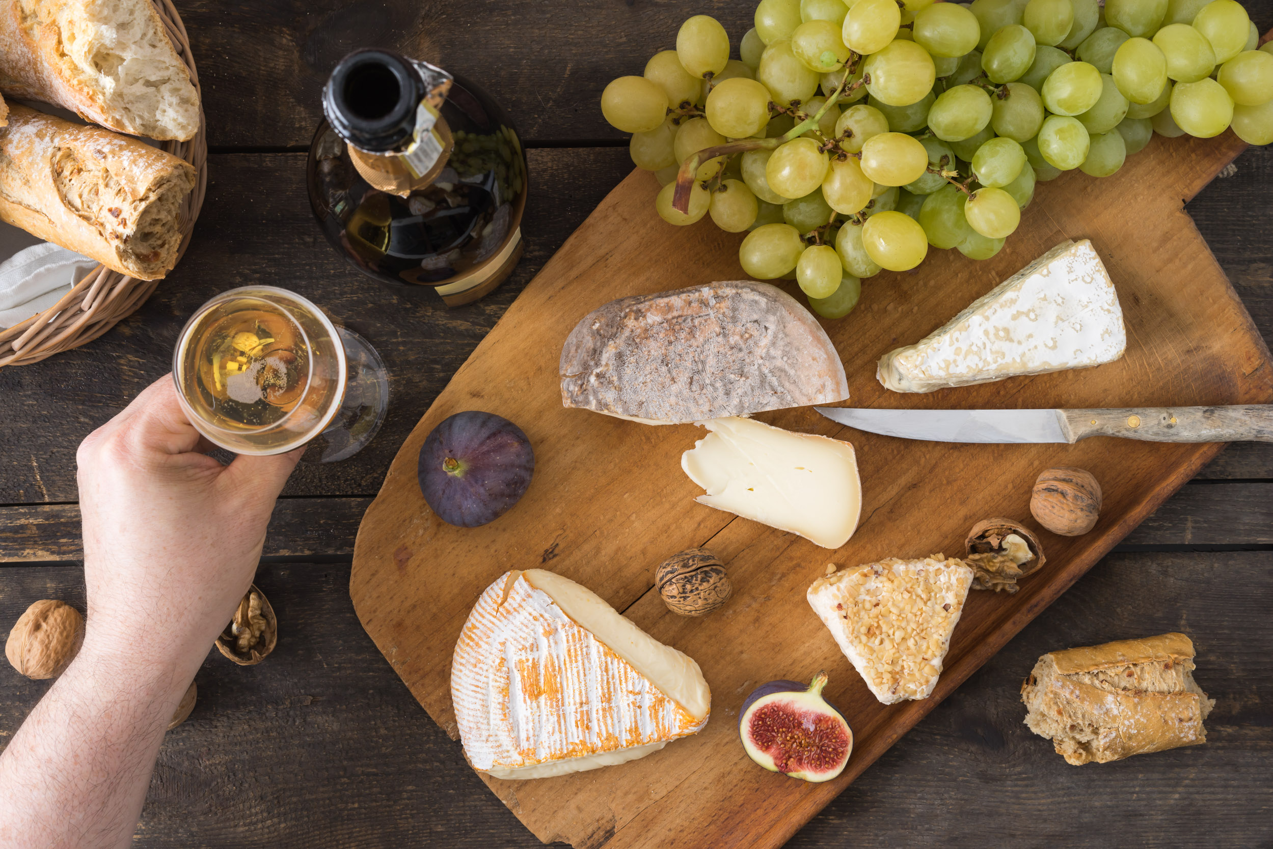Hand greift zum Champagner-Glas. Käseplatte mit Camenbert, Walnusskäse, Gorgonzola, Taleggio, dazu Champagner, Weintrauben, Feigen, Nüsse und Baguette