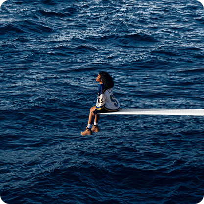SZA sitting on a boat's edge with legs hanging over the water, looking out at the ocean.