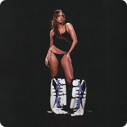 Tate McRae standing on a black background, wearing a black bra and panties with hockey goalie leg pads.