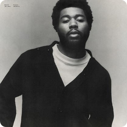 Black and white portrait of Khalid with a beard and natural hair, wearing a dark jacket over a light shirt, posed with a serious expression.