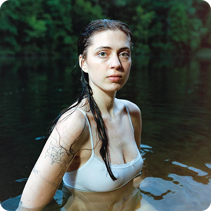 Lizzy McAlpine with wet hair wearing a white tank top in a lake surrounded by trees.